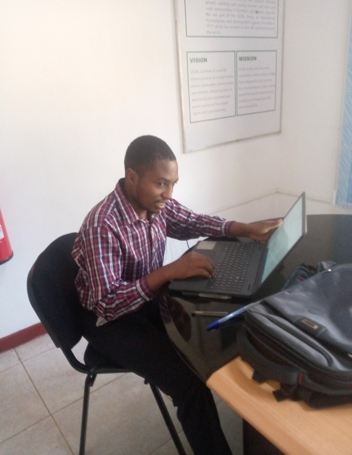 MR. Akankwatsa seated on a chair, a table with a laptop which he is using
